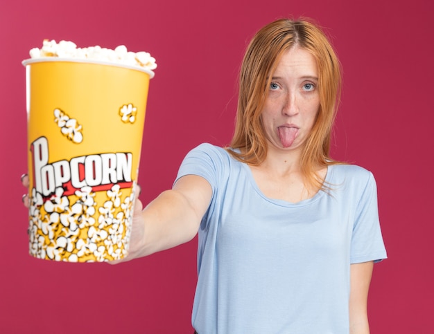 Una giovane ragazza rossa e rossa delusa con le lentiggini tira fuori la lingua e tiene un secchio di popcorn