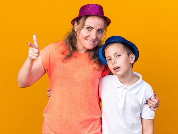 disappointed young slavic boy with blue party hat standing with his mother wearing purple party hat and pointing at side isolated on orange wall with copy space