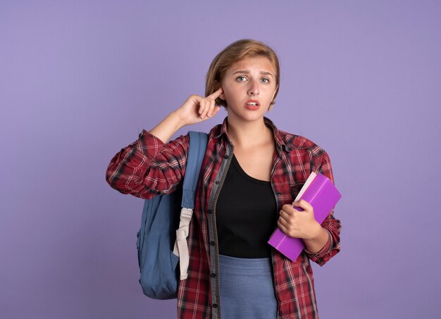 Disappointed young slavic student girl wearing backpack holds book and notebook closes ear with finger 