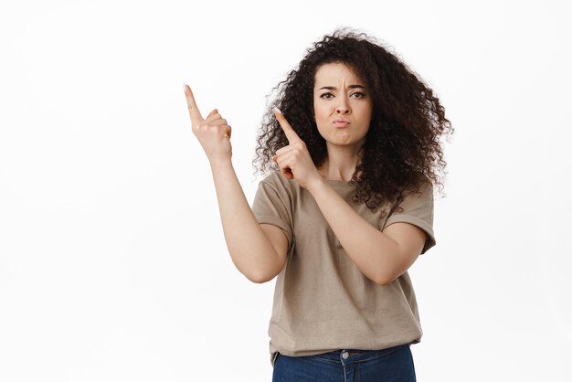 Disappointed young woman pointing at direction showing logo at upper left corner with grimacing upset frowning displeased complaining standing over white background