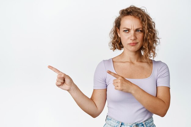 Disappointed young woman with short curly hair, frowning and sulking, pointing fingers left with dislike face on white