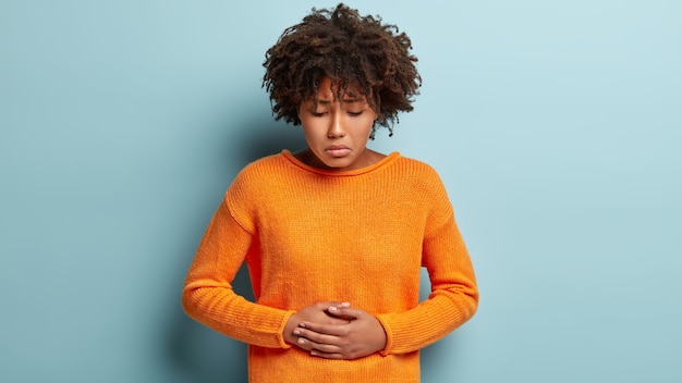 無料写真 不満アフロアメリカ人女性は腹痛に苦しんでおり、両手で腹に触れ、見下ろし、オレンジ色のジャンパーを着て、月経があり、巻き毛のヘアカットがあり、青い壁にモデルがいます。