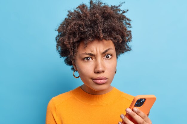 Discontent Afro American woman smirks face looks with dissatisfied expression holds modern cellular reads negative news online wears orange jumper isolated over blue wall. Technology concet