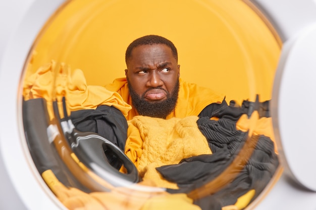 Discontent bearded man looks unhappily away smirks face sticks through laundry with liquid powder looks away poses from inside of washig machine in laundromat