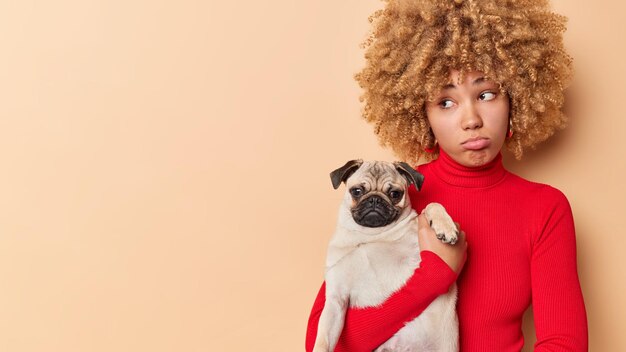 Discontent curly haired European woman purses lips looks sadly away poses with pug dog feels lonely lives alone with pet isolated over brown background copy space for your advertising content