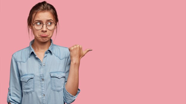 Discontent European woman with dejected facial expression, purses lower lip, points aside with thumb