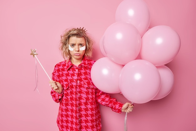 Free photo discontent fair haired woman prepares for costume party looks offended has spoiled makeup applies white beauty patches under eyes to reduce wrinkles holds bunch of inflated balloons stands indoor