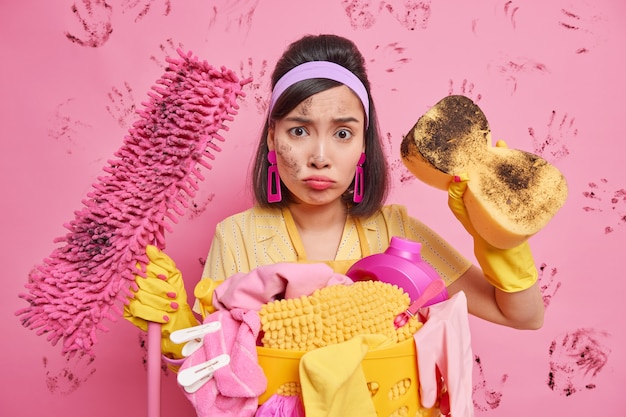 Discontent housewife looks sadly at camera has dirty face holds cleaning equipment poses near laundry basket with clothespins isolated over pink wall