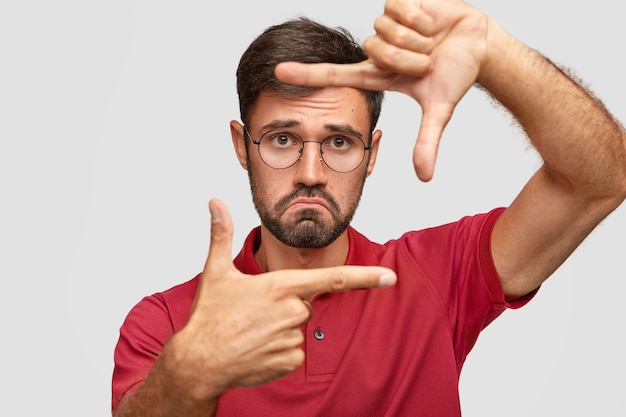 Discontent male movie director holds hands in framing gesture, look through with unhappy expression, wears round spectacles and red t-shirt, sees something unpleasant, isolated over white wall