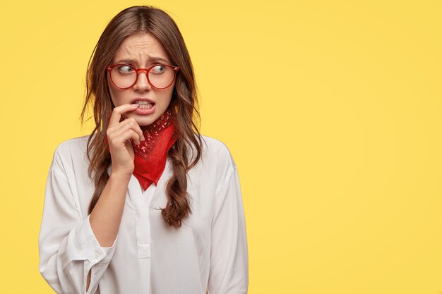 Discontent puzzled girl looks anxiously with worried expression aside, keeps fore finger near mouth, afraids of something, wears red handkerchief on neck and white shirt, copy space for advertisement