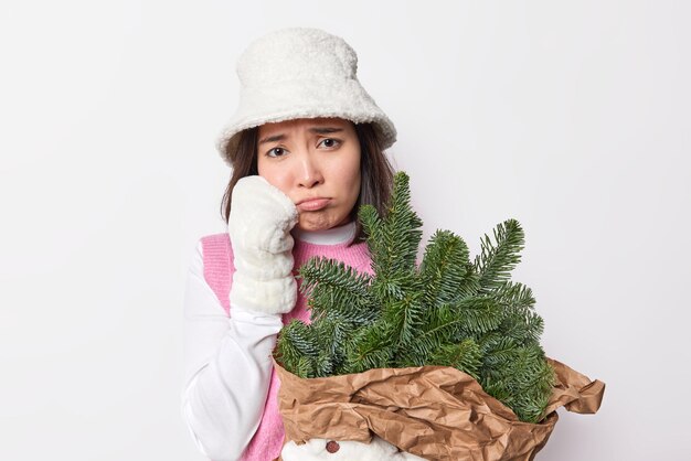 Free photo discontent sad woman has bad mood feels lonely during winter holidays looks unhappily dressed in white panama and mittens holds bouquet of green fir tree branches poses indoor. new year is coming