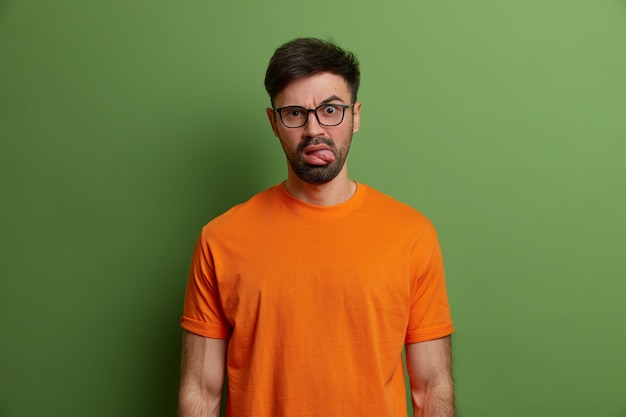 Discontent unshaven young European man sticks out tongue, makes bothered displeased face expression, wears transparent glasses and bright orange t shirt, isolated over bright green wall