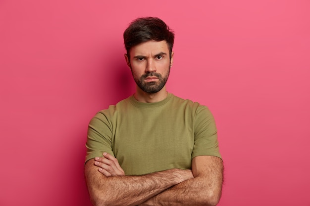 Foto gratuita il giovane malcontento con la barba lunga aggrotta le sopracciglia, guarda con espressione seria e rigorosa, tiene le mani incrociate sul corpo, ascolta qualcuno, aspetta spiegazioni, chiede verità, indossa una maglietta casual