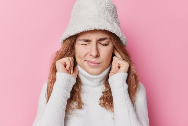 不満のある女性は、大きな音や隣人からの騒音に悩まされている指で耳を塞ぎ、目を閉じたまま、ピンクの背景の上に白い帽子とセーターを着ています。もう聞こえない