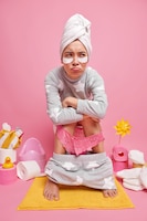 Discontent young asian woman suffers from constipation or hemorrhoids poses on toilet bowl applies beauty patches under eyes dressed in soft pajama has stomachache isolated over pink wall