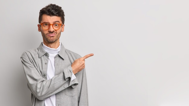 Discontent young man expresses aversion and dislike points index finger aside on copy space disappointed with bad product dressed in stylish clothes isolated over grey wall. Disgusting thing