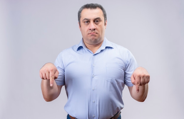 Free photo discontented middle age man wearing blue vertical stripped shirt showing index fingers downward while standing