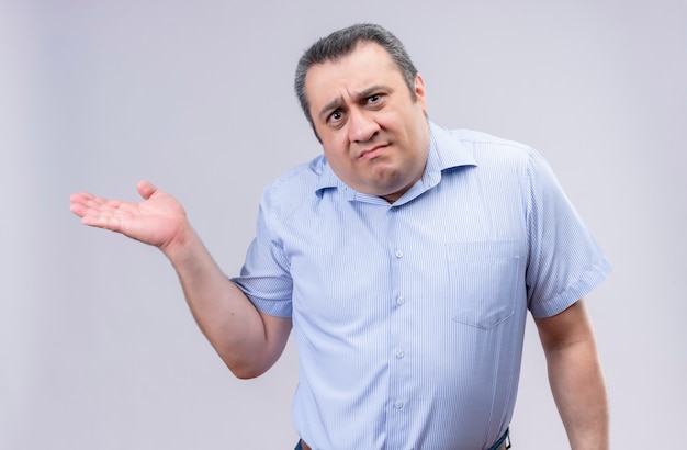 Discontented middle-aged man wearing blue vertical stripes shirt spreading his hand while standing 