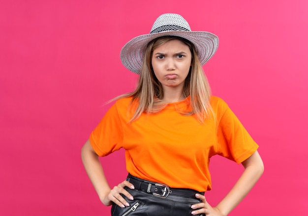 A discontented pretty young woman in an orange t-shirt wearing sunhat keeping hands on waist while looking on a pink wall