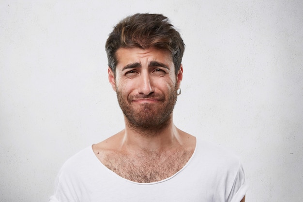 Discouraged man with hairdo and beard frowning his face being sorry for what he done. Grieved man in white T-shirt . People, fashion, lifestyle, emotions concept