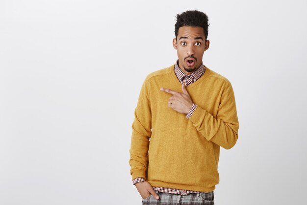 Discussing with friend shocking gossip about classmate. Attractive african-american male student with afro hairstyle in yellow pullover, dropping jaw from surprise, pointing at upper left corner