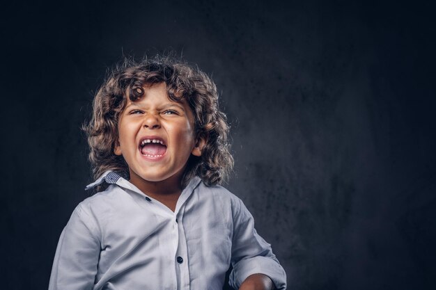 Disgruntled schoolboy with brown curly hair dressed in a white, cry at a studio. Isolated on a dark textured background.