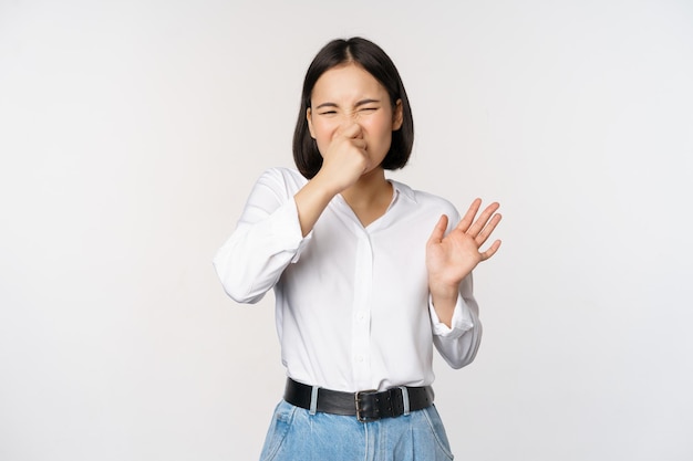 Free photo disgusted asian girl close nose and showing rejection gesture digusting bad smell standing over white background