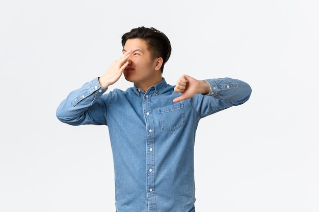 역겨운 아시아 남자는 손가락으로 코를 막고 엄지손가락을 아래로 내밀고 지독한 악취를 호소하고 악취를 풍기며 나쁜 냄새에 시달리고 흰색 배경에 서 있습니다.