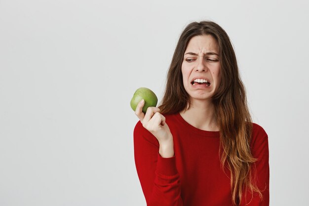 Disgusted cute woman looking at apple with aversion and dislike, grimacing