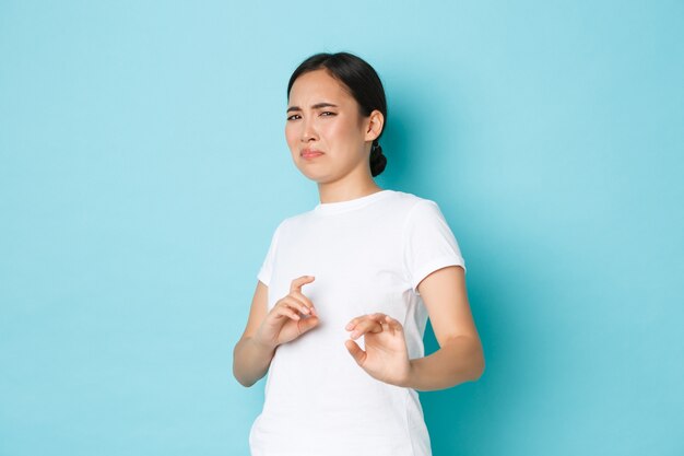 Disgusted and displeased asian girl expressing aversion and reluctance, grimacing and raising hands defensive, stay away, refusing offer, rejecting something, standing blue wall