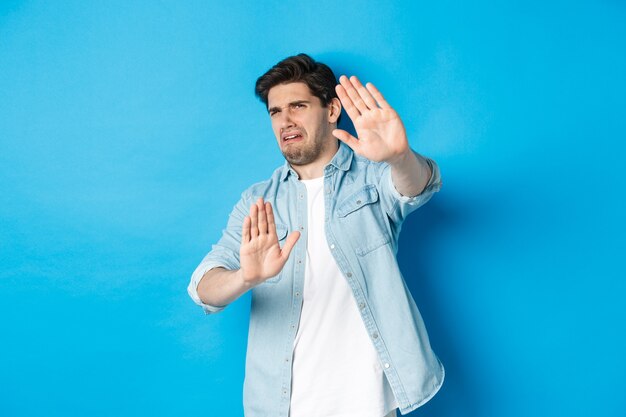 Disgusted guy saying no, refusing and looking away from something awful, cringe while standing against blue background