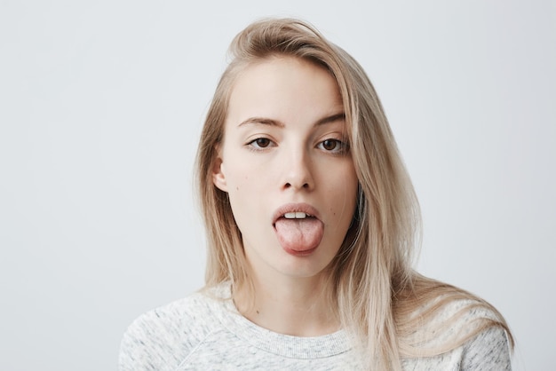 Disgusted pretty young woman with blonde hair sticking out tongue expressing her dislike or disregard towards something Funny humorous female model showing tongue at camera as if teasing someone