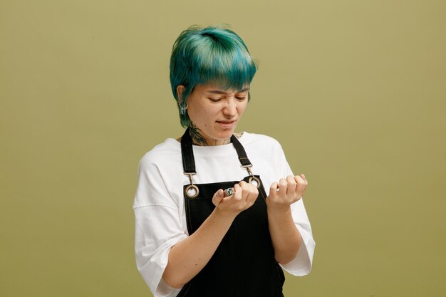 Disgusted young female barber wearing uniform holding nail clippers looking at her nails isolated on olive green background