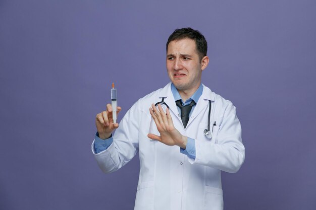 Disgusted young male doctor wearing medical robe and stethoscope around neck holding syringe with needle looking at it showing no gesture isolated on purple background