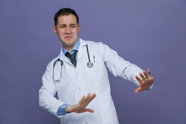Disgusted young male doctor wearing medical robe and stethoscope around neck looking at side making no gesture isolated on purple background