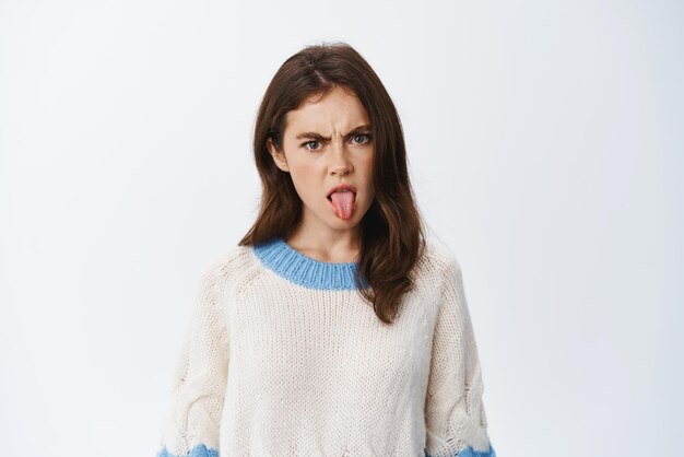 嫌な若い女性が眉をひそめ、白い背景のセーターに立っている悪い味に不快な何かのひどいまたは嫌なことに舌を見せている