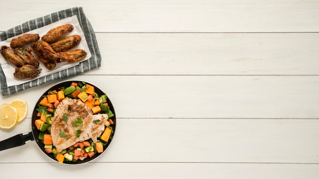 Блюдо с куриными крылышками и сковородой овощей на деревянный стол