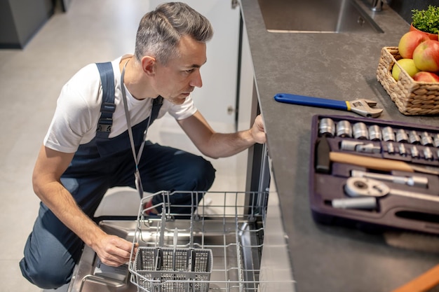 Dishwasher repair Top view of middleaged man crouching near open dishwasher