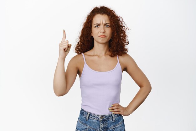 Dislike. Portrait of young redhead curly woman frowning, pointing finger up and furrow eyebrows displeased, dislike smth, standing over white background