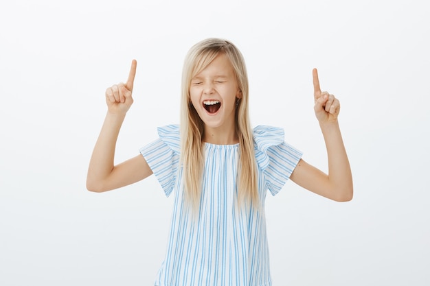 Free photo disobedient energetic little blond girl, raising index fingers and pointing up, yelling or screaming out loud with closed eyes, being disobedient while demanding buy new toys, standing over gray wall