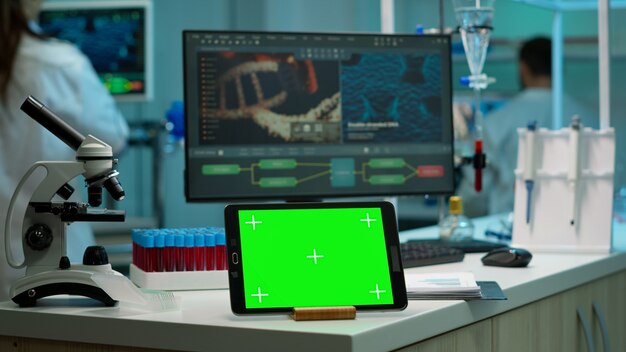 緑色の画面でタブレットを表示し、科学実験室の机の上に置かれたテンプレートにモックアップし、女性の医学研究者が実験を行うデジタルモニターでウイルスの進化を分析している