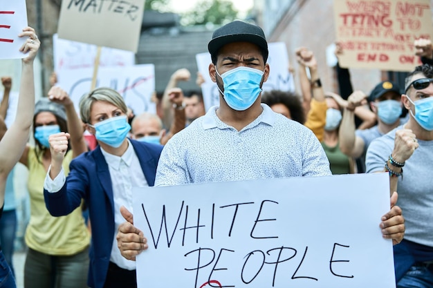Displeased African American man wearing protective face mask and carrying a placard while protesting with crowd of people on the streets