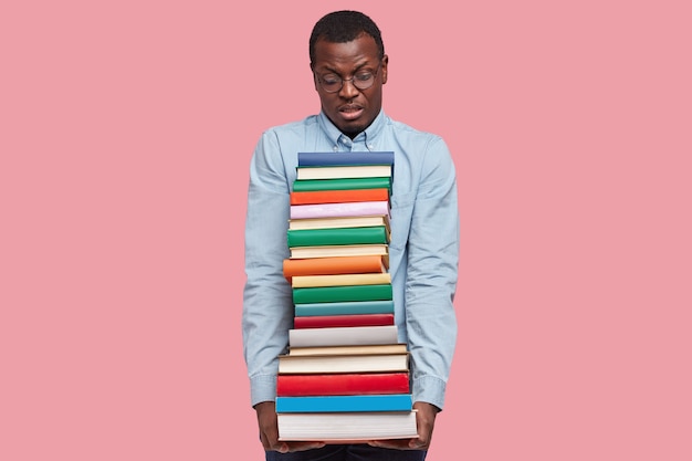 Free photo displeased african american scientist or student carries heavy pile of textbooks, wears elegant shirt