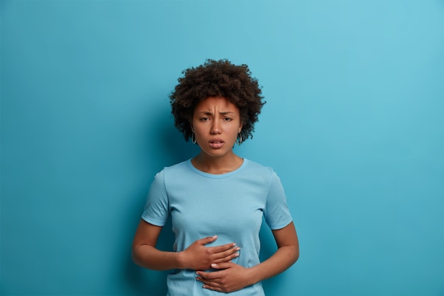 La donna afro scontenta avverte fastidio allo stomaco, tiene i palmi sulla pancia, ha crampi mestruali, indossa una maglietta casual, mangia cibo avariato, isolata sul muro blu, esprime emozioni negative con la mimica