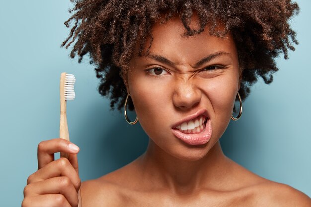 不機嫌なアフロの女性は顔を眉をひそめ、歯を食いしばり、口腔衛生の世話をし、巻き毛を持ち、歯ブラシを持ち、裸の肩を示し、青い壁の上でポーズをとります。