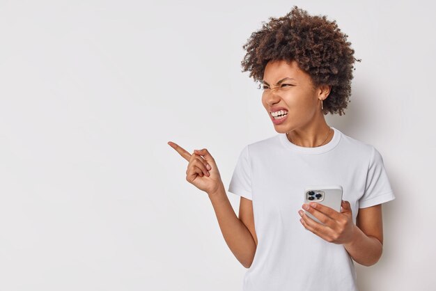 Displeased angry woman looks with irritated expression away points index finger on blank space shows place for your advertisement holds mobile phone dressed casually isolated over white wall