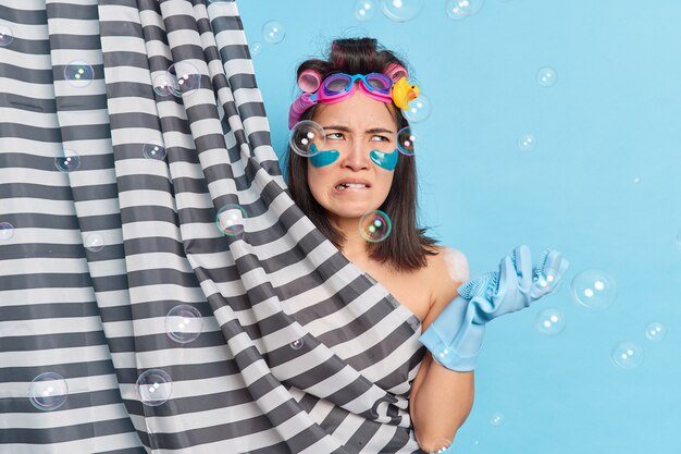 Displeased annoyed young Asian woman undergoes hygiene and beauty treatments wears swimming goggles rubber gloves hides naked body behind shower curtain 