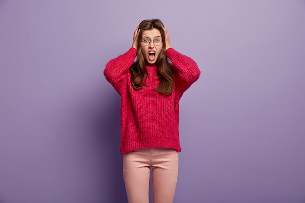 Displeased annoyed young woman shouts angrily, keeps hands on head, expresses negative emotions, wears round glasses, red jumper, irritated with something bad, isolated over purple wall