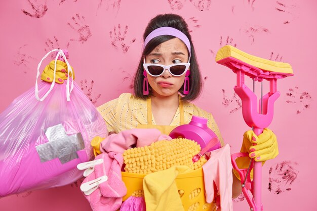 Displeased Asian lady cleans messy room after party looks sadly at trash bag looks tired while cleaning home holds mop wears trendy sunglasses poses near laundry basket against dirty wall