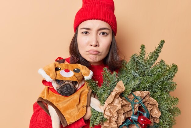 Displeased Asian millennial girl looks sadly at camera holds pedigree dog in winter clothes fir tree branchess wrapped in paper and retro garland wears red hat isolated over beige background